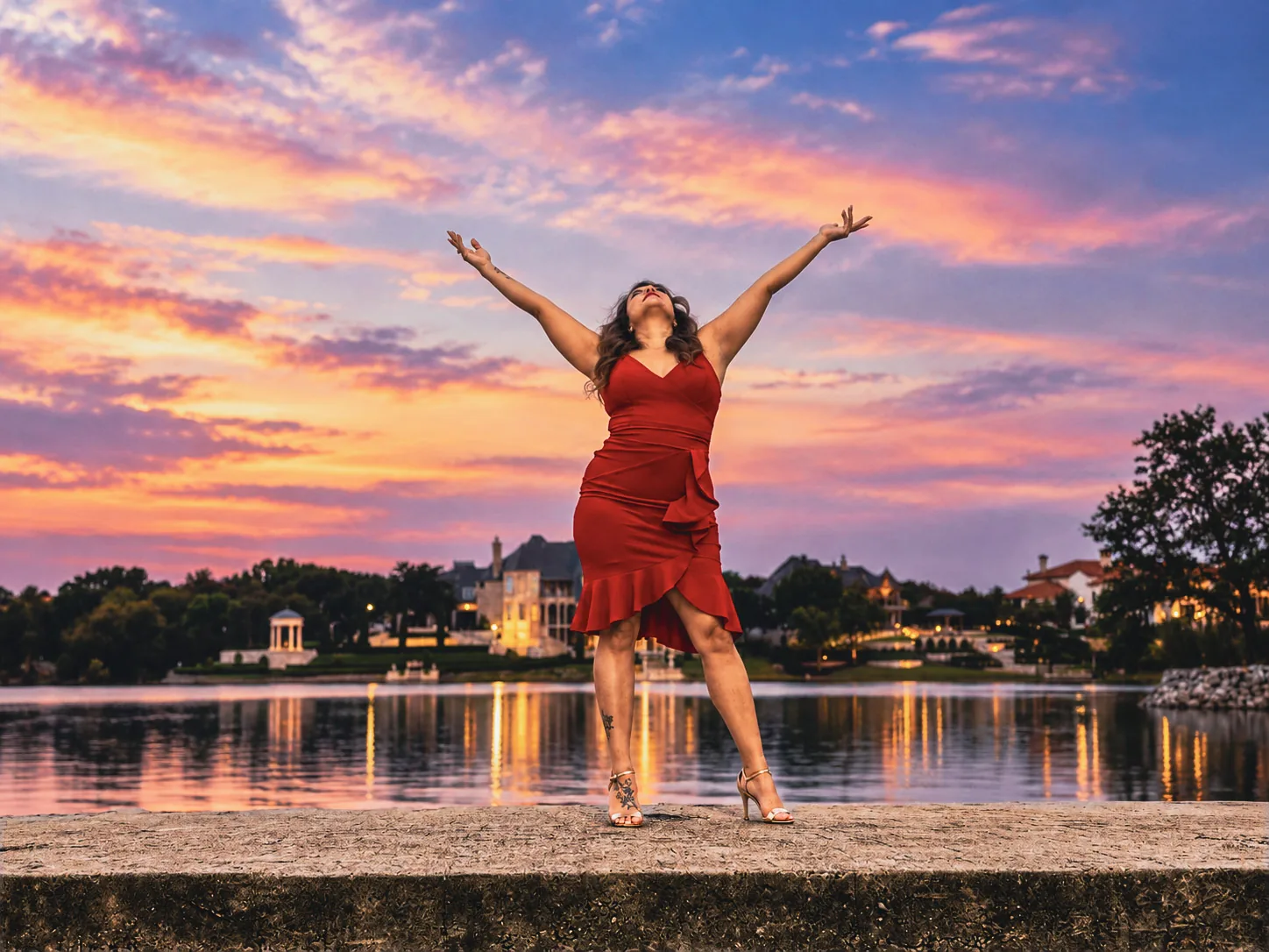 A woman in a red dress posing beside the water at sunset.